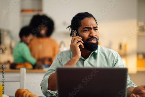A serious african american casual businessman is sitting at the breakfast table with a laptop at home and having an important phone call while his family preparing breakfast in the kitchen.