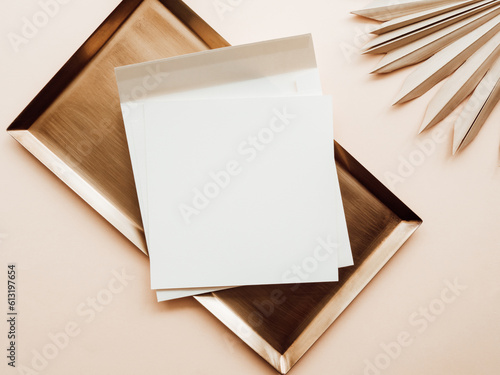 White blank paper square card mockup, envelope on gold tray and dry palm leaf on beige background
