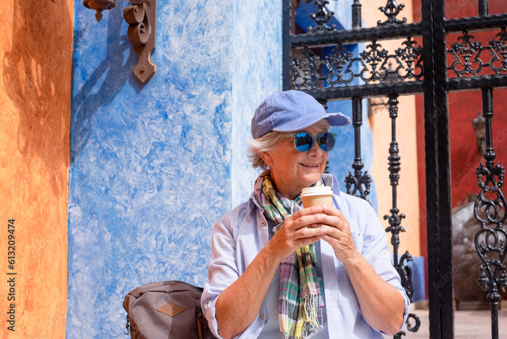 Happy senior woman in leisure travel sitting on a staircase having a coffee break. Active elderly lady casual clothing with sunglasses and cap enjoying vacations