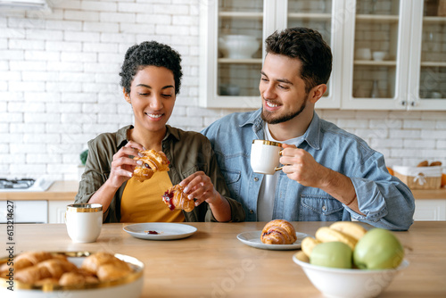 Morning breakfast. Happy multiracial couple, caucasian man and african american woman, spend time together at home, they drink coffee, eat croissants, chat on different topics, plan leisure time,smile