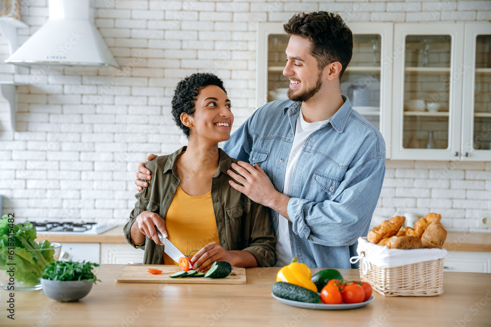 Multiracial happy couple in casual wear, young spouses, stand at home in the kitchen, prepare salad together, woman cuts vegetables, man hugs her, they look at each other with love, smile Multiracial happy couple in casual wear, young spouses, stand at home in the kitchen, prepare salad together, woman cuts vegetables, man hugs her, they look at each other with love, smile