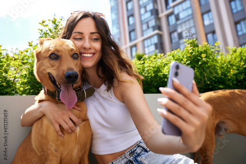 A charming young smiling girl is resting on a bench with two golden-colored dogs during a walk on a sunny day. The girl plays with pets and takes a selfie. Love between owner and pet. photo