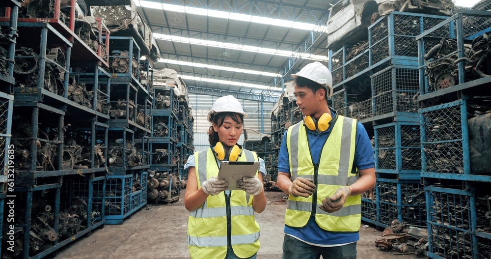 Two asian warehouse workers wearing safety helmet holding tablet while walking at automotive parts warehouse for checking stock. Vehicle part, Recycling Two asian warehouse workers wearing safety helmet holding tablet while walking at automotive parts warehouse for checking stock. Vehicle part, Recycling