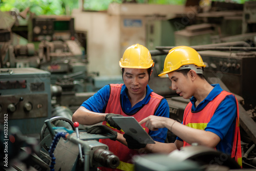 Two young asian male technicians wearing safety uniform workwear, helmets, vest and gloves are Working on a metal lathe and inspecting parts intently In industrial plants.