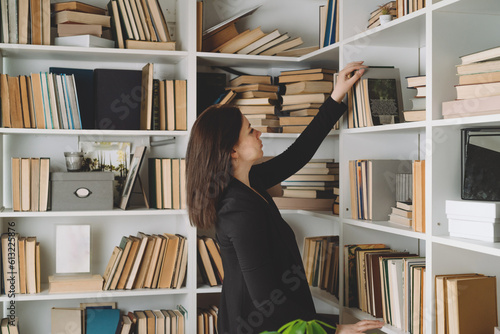 Young woman chooses a book in a bookcase at home. Woman looks through the books in the library, deciding which one to take to study. Book Lovers Day. Copy space