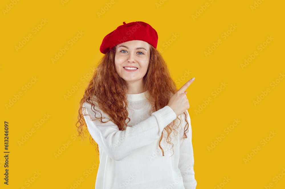 Portrait of smiling, happy young girl in red beret wearing white sweater points her finger to the side. Studio photo of beautiful girl with long wavy red hair on isolated yellow background.