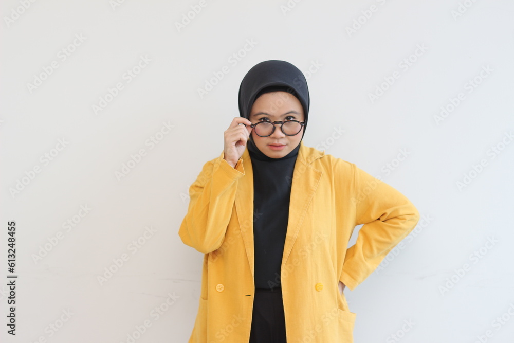 Young beautiful Asian Muslim woman, wearing yellow blazer looking at camera holding glasses curiously isolated on white background. Young beautiful Asian Muslim woman, wearing yellow blazer looking at camera holding glasses curiously isolated on white background.