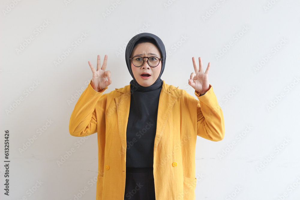 beautiful young Asian Muslim woman, wearing glasses and yellow blazer with hand gesture of approval or OK beautiful young Asian Muslim woman, wearing glasses and yellow blazer with hand gesture of approval or OK