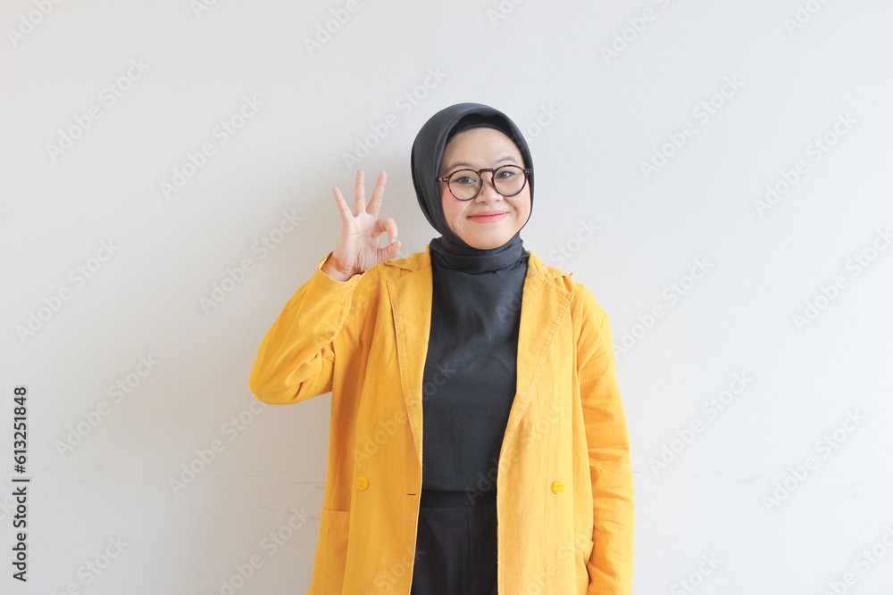 beautiful young Asian Muslim woman, wearing glasses and yellow blazer with hand gesture of approval or OK beautiful young Asian Muslim woman, wearing glasses and yellow blazer with hand gesture of approval or OK