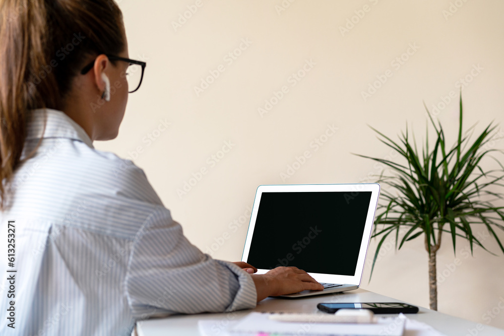 Rear view businesswoman wears glasses and wireless earphones working on her laptop in the office, blank screen of laptop. Rear view businesswoman wears glasses and wireless earphones working on her laptop in the office, blank screen of laptop.