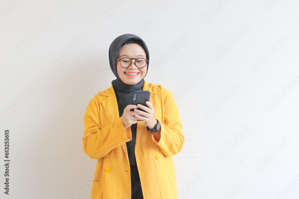 beautiful young asian woman in glasses, hijab and wearing a yellow blazer looking at her mobile phone with happy face beautiful young asian woman in glasses, hijab and wearing a yellow blazer looking at her mobile phone with happy face