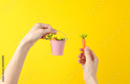 Hands holding a plastic miniature carrot and backet on yellow background. Gardening photo