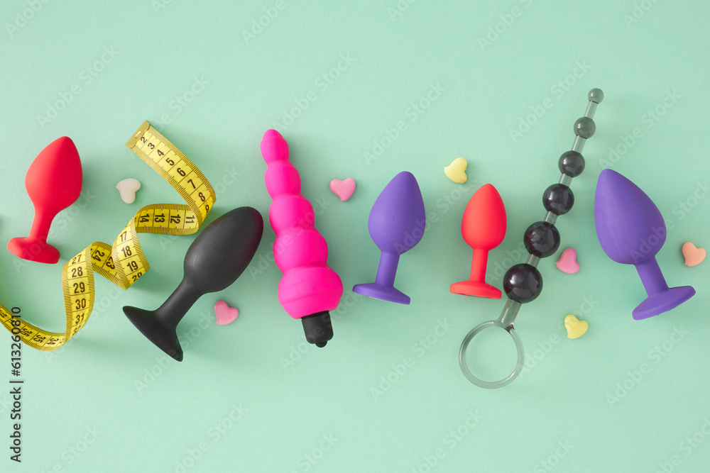 Concept of size options in adult sex toy selection. Top view arrangement different anal butt plugs and anal beads measuring tape and colorful hearts on turquoise background.