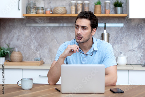 Portrait of handsome man sitting at the table and working on laptop in the kitchen at home 