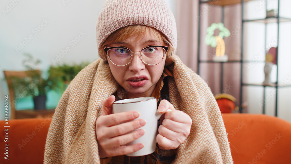 Sick young woman wear hat wrapped in plaid sit alone shivering from cold on sofa drinking hot tea in unheated apartment. Unhealthy ill girl in glasses feeling discomfort try to warming up at home room