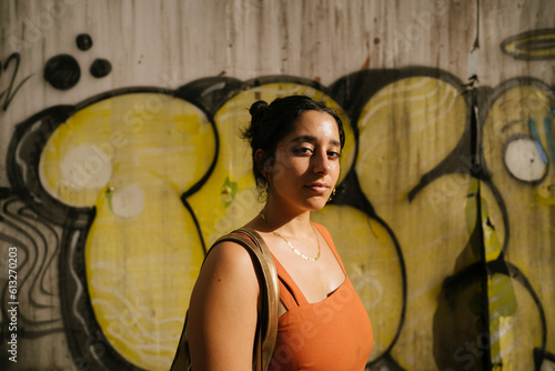 medium portrait of a pretty latina woman on an orange dress facing the sun during golden hour in front of graffiti (ID: 613270203)