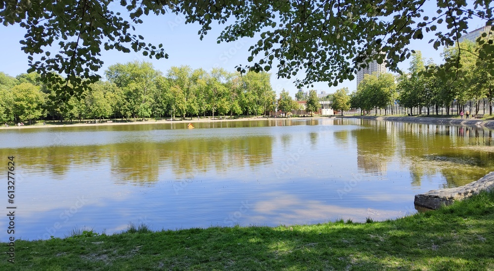 Lake in city public square. Beautiful water landscape. Park by Becket Pond in Moscow. Sunny summer day. Trees and sky are reflected in water of reservoir. Nature for recreation in middle of metropolis Lake in city public square. Beautiful water landscape. Park by Becket Pond in Moscow. Sunny summer day. Trees and sky are reflected in water of reservoir. Nature for recreation in middle of metropolis