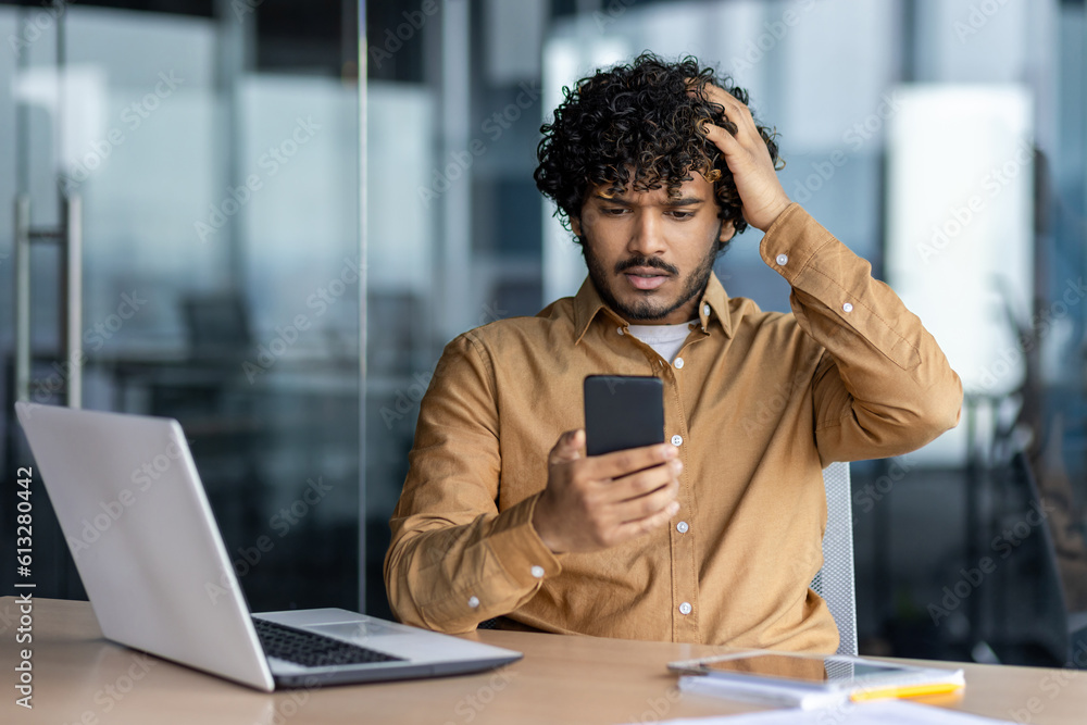 Upset and sad man at workplace inside office, businessman at work received notification message negative, hispanic reading bad news online from smartphone.