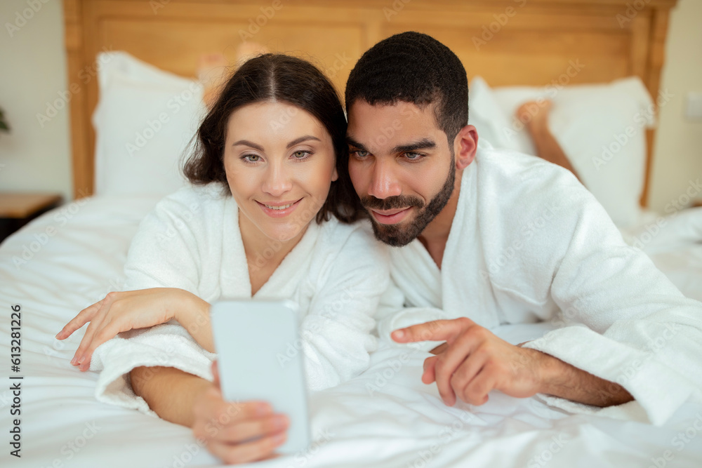Cheerful Couple Using Smartphone Lying In Bed In Hotel Indoors Cheerful Couple Using Smartphone Lying In Bed In Hotel Indoors