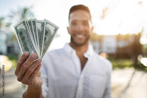Happy young businessman holding fan of dollar money and smiling at camera, standing outdoors, selective focus, closeup
