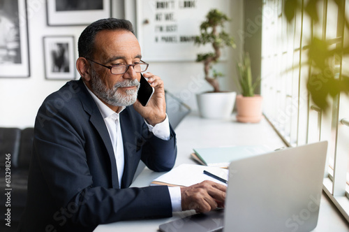 Smiling european senior businessman in suit, glasses with laptop calls by smartphone at table in office