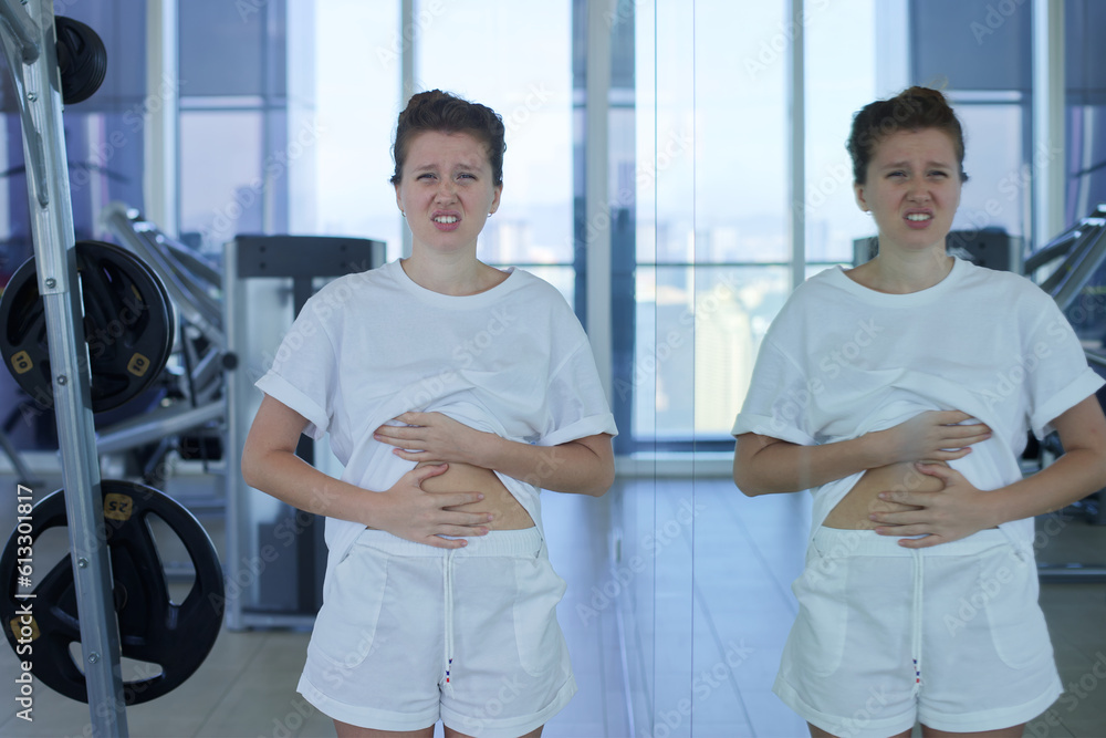 Fototapeta premium Young sad upset woman is disappointed because of fat belly, stomach, looking at mirror with frustrated look, trying to loose weight 