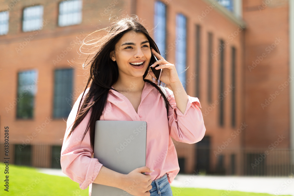 Fototapeta premium Excited hispanic lady making phone call, talking on cellphone while walking in college campus after classes, free space