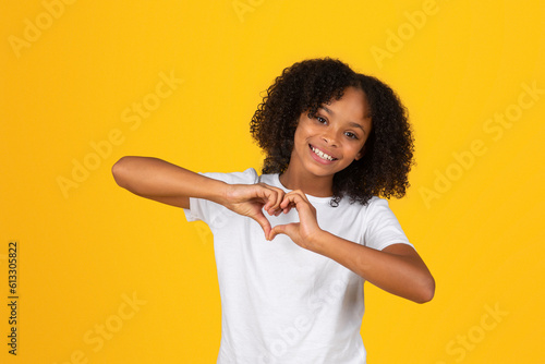 Positive curly teen black schoolgirl in white t-shirt has fun, making heart gesture with hands