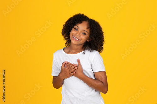 Satisfied glad curly teenager black girl in white t-shirt presses hands to chest, says thank