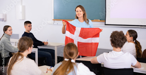 Adult female teacher in the classroom showing the flag of Denmark to the students at the geography lesson