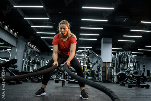 Attractive young fit female working out at the gym. She's doing exercise with battle ropes. 