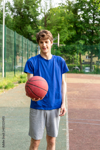 Cute boy in t shirt plays basketball on city playground. Active teen enjoying outdoor game with red ball. Hobby, active lifestyle, sport for kids.  © Natali