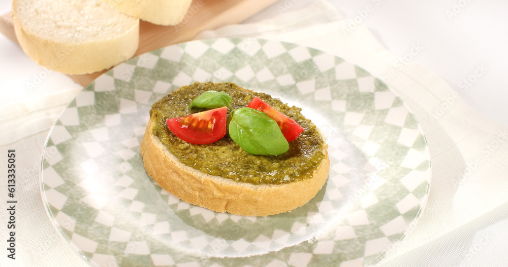 bread with pesto, basil and tomato on a plate. slices of bread on a white tablecloth. traditional italian green sauce. angle view. sunny day. bread with pesto, basil and tomato on a plate. slices of bread on a white tablecloth. traditional italian green sauce. angle view. sunny day.