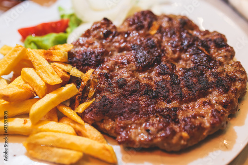 Pljeskavica with french fries on a plate, traditional serbian dish, grilled dish of spiced meat patty mixture of pork, beef and lamb, popular balkan steet food Pleskavitza, served in cafe photo