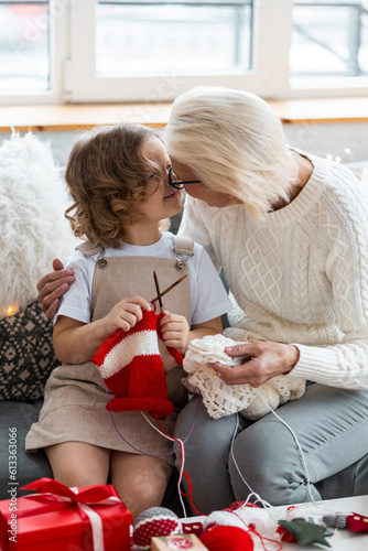 Grandmother and granddaughter spend quality time together doing craft toys and knitting near decorated Christmas new year tree. Cute little girl and attractive senior woman at home in the living room