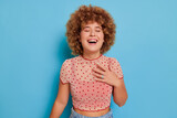 Happy cute curly girl in pink polka-dot top standing pressing hand to heart and laughing with eyes closed, fun time concept, copy space