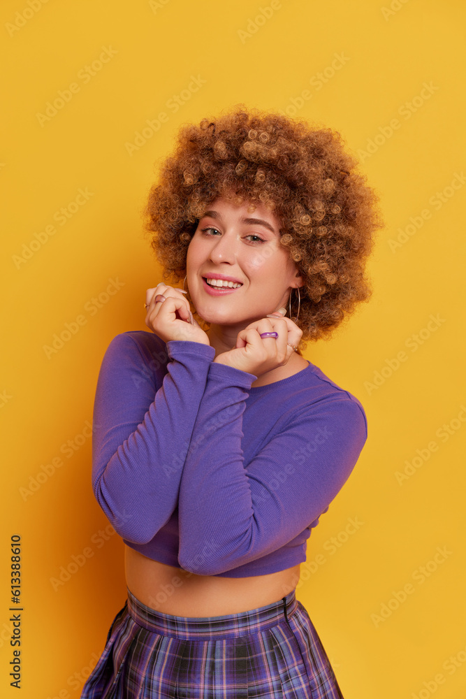 Beautiful girl in curly wig dressed in purple top and plaid skirt posing on yellow background, pressing her hands to her chest, funny moments concept, copy space Beautiful girl in curly wig dressed in purple top and plaid skirt posing on yellow background, pressing her hands to her chest, funny moments concept, copy space