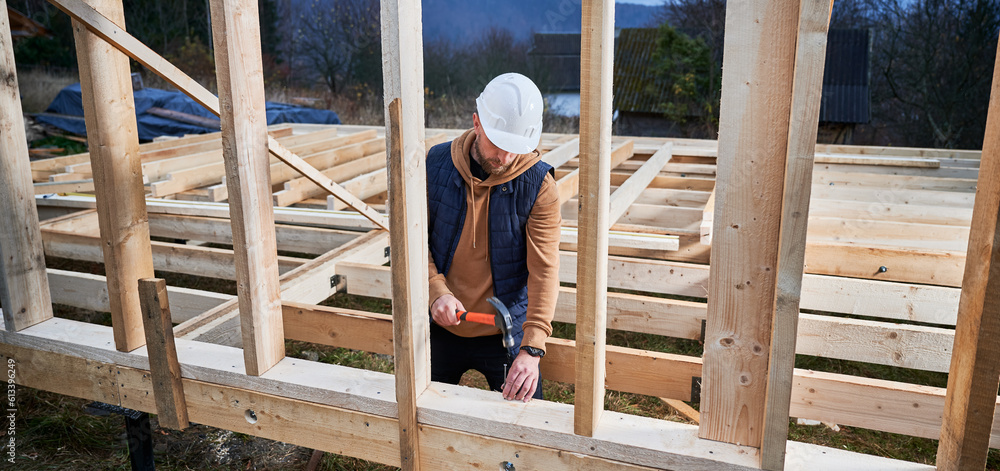 Man worker building wooden frame house on pile foundation. Carpenter hammering nail into wooden plank, using hammer. Carpentry concept. Man worker building wooden frame house on pile foundation. Carpenter hammering nail into wooden plank, using hammer. Carpentry concept.