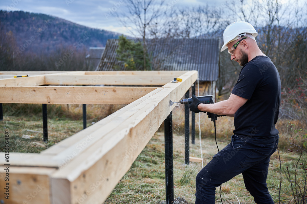 Man building wooden frame house on pile foundation. Male worker drilling hole by drill in timber framing of future house. Builder wearing white helmet and protective goggles. Carpentry concept. Man building wooden frame house on pile foundation. Male worker drilling hole by drill in timber framing of future house. Builder wearing white helmet and protective goggles. Carpentry concept.