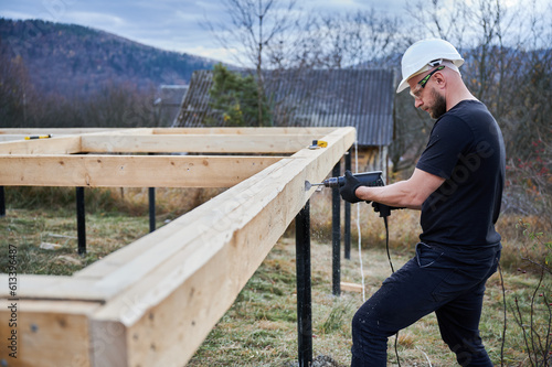 Man building wooden frame house on pile foundation. Male worker drilling hole by drill in timber framing of future house. Builder wearing white helmet and protective goggles. Carpentry concept.