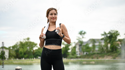 A beautiful Asian woman in sportswear stands with a towel on her neck in the public park.