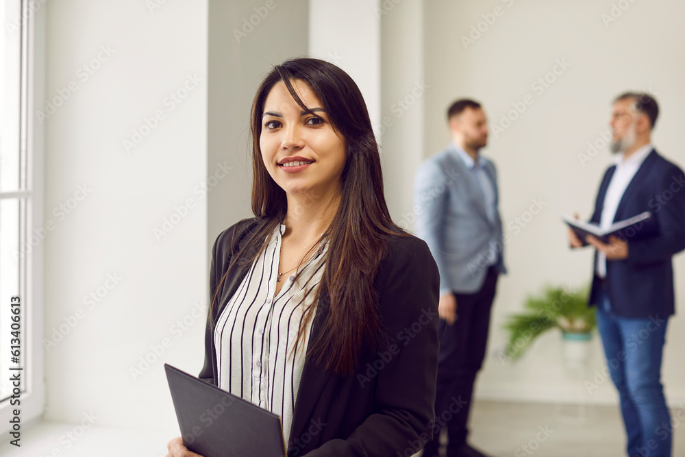 Successful businesswoman standing in office on background of colleagues. Smiling confident young woman in formal wear holding clipboard, posing in modern office, looking at camera Successful businesswoman standing in office on background of colleagues. Smiling confident young woman in formal wear holding clipboard, posing in modern office, looking at camera