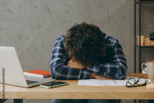 Sad tired exhausted sleepless employee business Indian man he wearing casual blue checkered shirt sleeping taking nap crying put head on table sit work at office desk with laptop pc computer indoors. photo