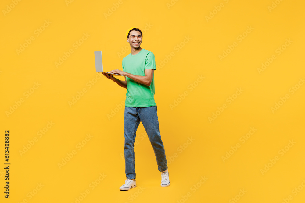 Full body young IT man of African American ethnicity he wear casual clothes green t-shirt hat hold use work on laptop pc computer isolated on plain yellow background studio portrait Lifestyle concept Full body young IT man of African American ethnicity he wear casual clothes green t-shirt hat hold use work on laptop pc computer isolated on plain yellow background studio portrait Lifestyle concept