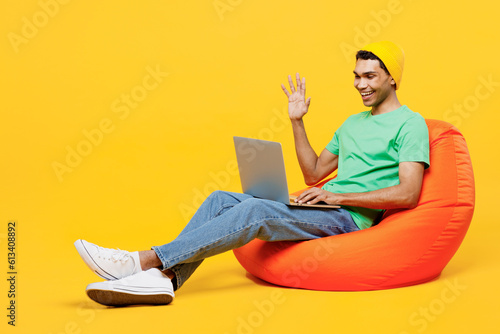 Full body young IT man of African American ethnicity wears casual clothes green t-shirt hat sit in bag chair hold use work on laptop pc computer waving hand isolated on plain yellow background studio.