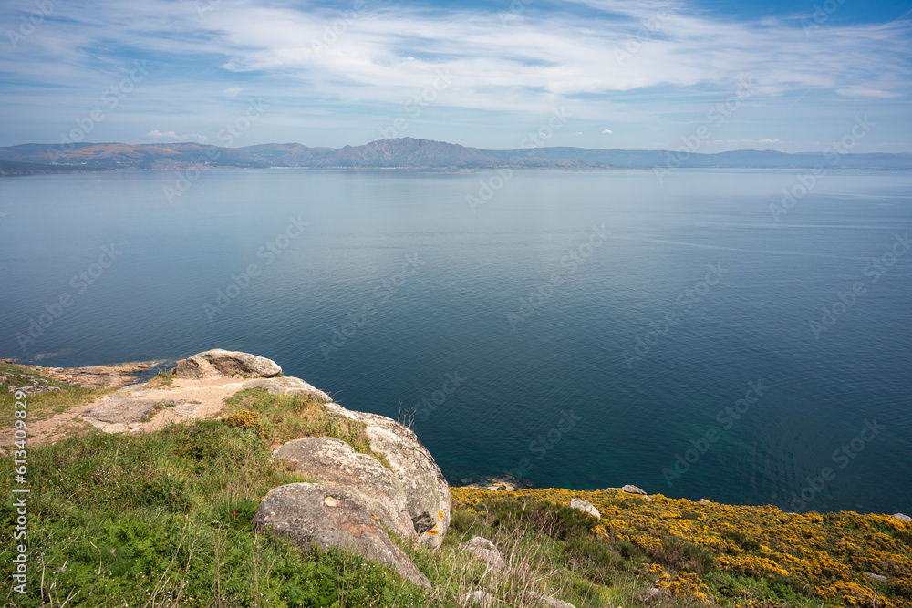 skyline image of a coastal seascape seen from the top of a grassy, grassy cliff where you can see the vast sea, the blue sky and the mountains in the distance on the other side.