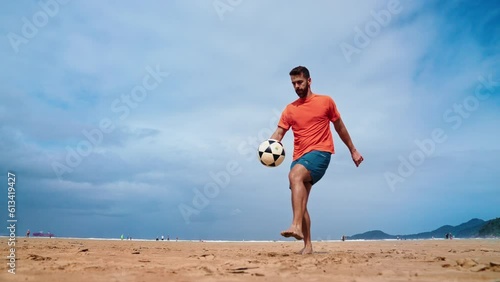 Football freestyle. Adult man practices with a soccer ball on the beach. Cinematic video of a adult man with a orange shirt playing football with the beach and the sea in the background.