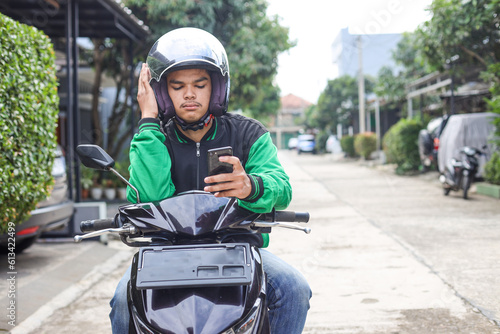 Asian online taxi driver wearing green jacket and helmet feeling bored while waiting order from smartphone