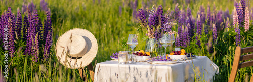 Sunset, golden hour. Romantic table decor for loving couple on blooming meadow with purple lupines. Two glasses of wine, flowers, silverware, fruits, wooden vintage furniture banner