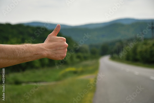 Outstretched hand and thumbs up. Rear view. Travel concept. Young man hitchhiker stands by rural road on sunny summer day. Only hand, no human's face. Guy went on trip alone, trying to stop car.